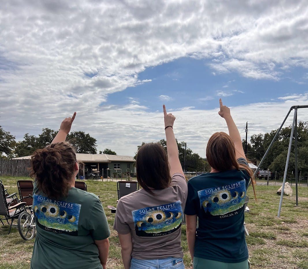 Three people in matching solar eclipse t-shirts point at the sky in a rural setting with scattered clouds.