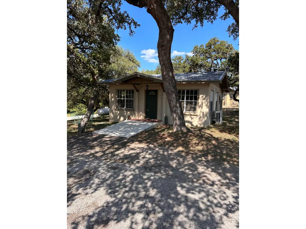 A small beige house with white trim, nestled under large oak trees casting shadows on the yard, against a clear sky.