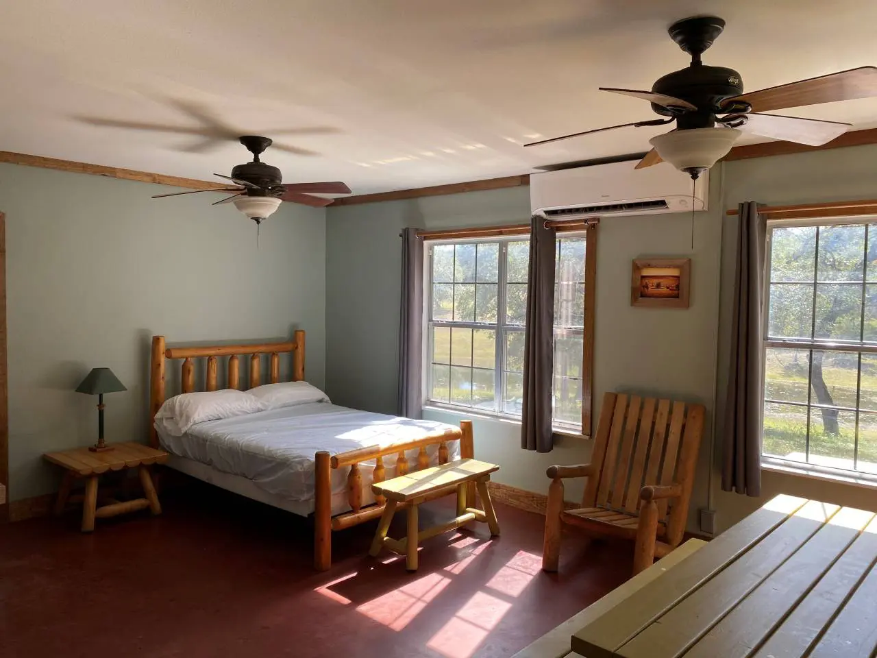 A sunny, simple bedroom with a single wooden bed, two ceiling fans, a rocking chair, and a stool beside a bed lamp. two windows let in natural light.