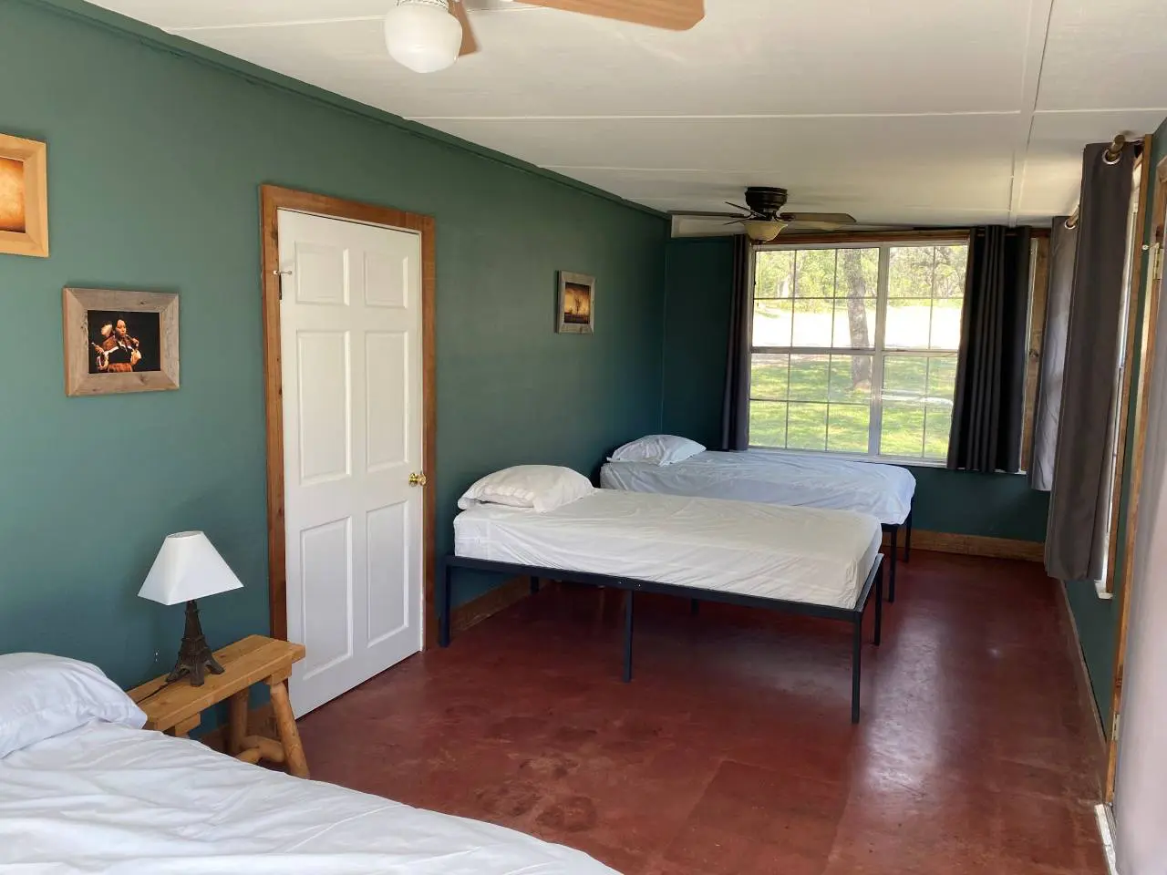 A simple bedroom with two twin beds, green walls, a white door, dark curtains, a ceiling fan, and a small lamp on a wooden nightstand.