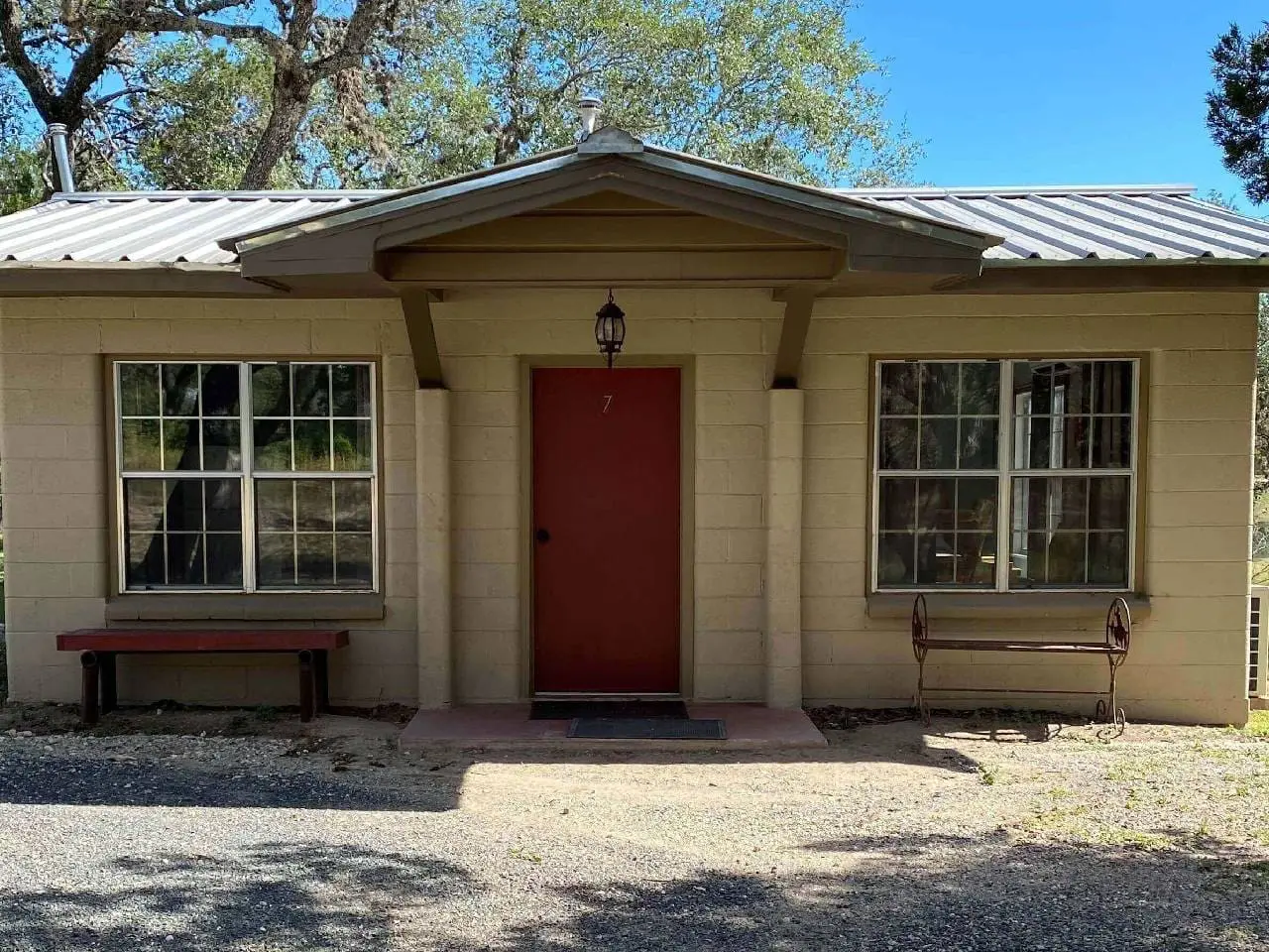 Small beige house with a red door, numbered 7, flanked by two benches under large windows, and a hanging lantern above the door.