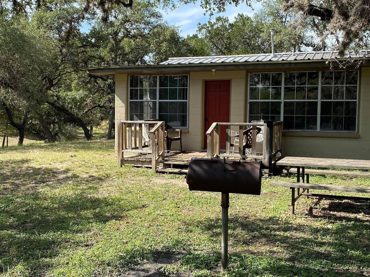 Rustic house with a red door, wooden porch, and a mailbox in the foreground surrounded by oak trees and grassy area.