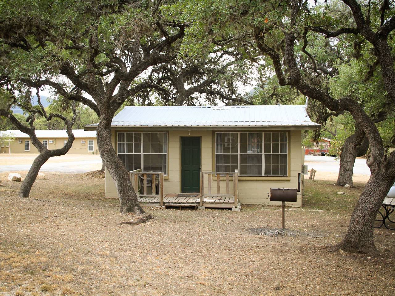 A small rustic cabin with a green door situated under sprawling oak trees, featuring a front porch and a mailbox, set in a tranquil park area.