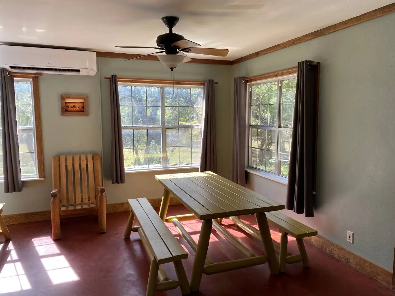 A bright, sunlit room with mint green walls featuring a wooden picnic table and bench, a rocking chair, and windows overlooking trees. a ceiling fan is above.