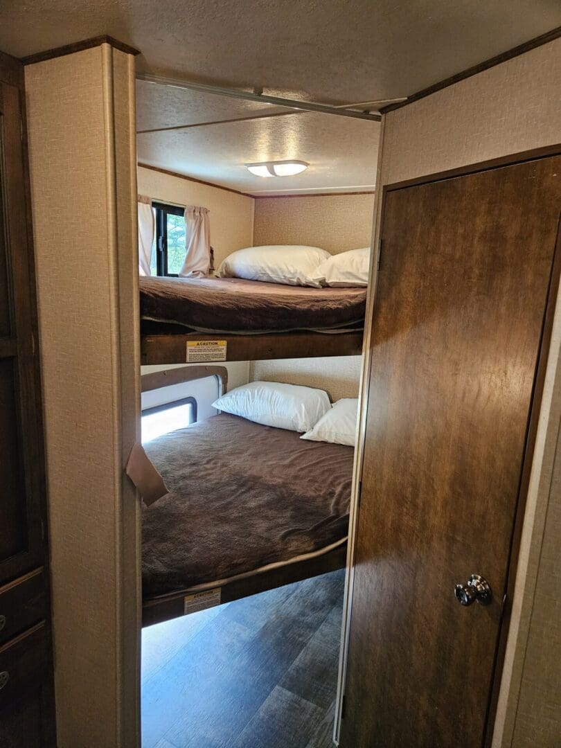 Interior of a camper with wooden bunk beds, showing a bottom bunk with brown bedding and an upper bunk with white bedding, alongside built-in wooden cabinets.