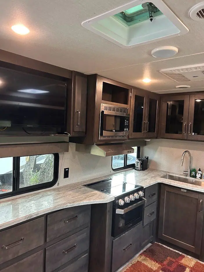 Interior of an rv showing a kitchen with dark wood cabinets, a microwave, stove, and a visible television.