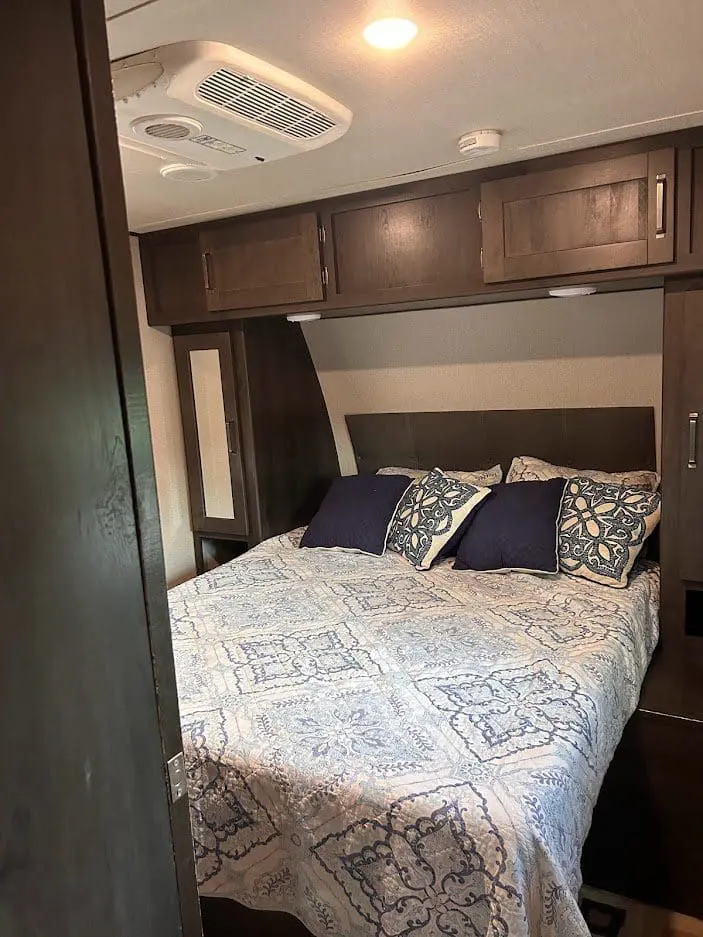 Interior of an rv bedroom featuring a bed with blue and white bedding and dark wood overhead cabinets.