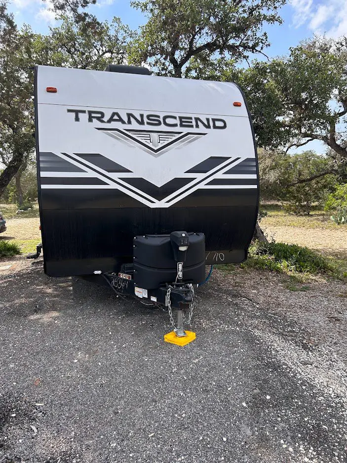 Front view of a parked "transcend" travel trailer featuring a distinctive black and white graphic design, equipped with a hitch and secured with a yellow wheel lock.