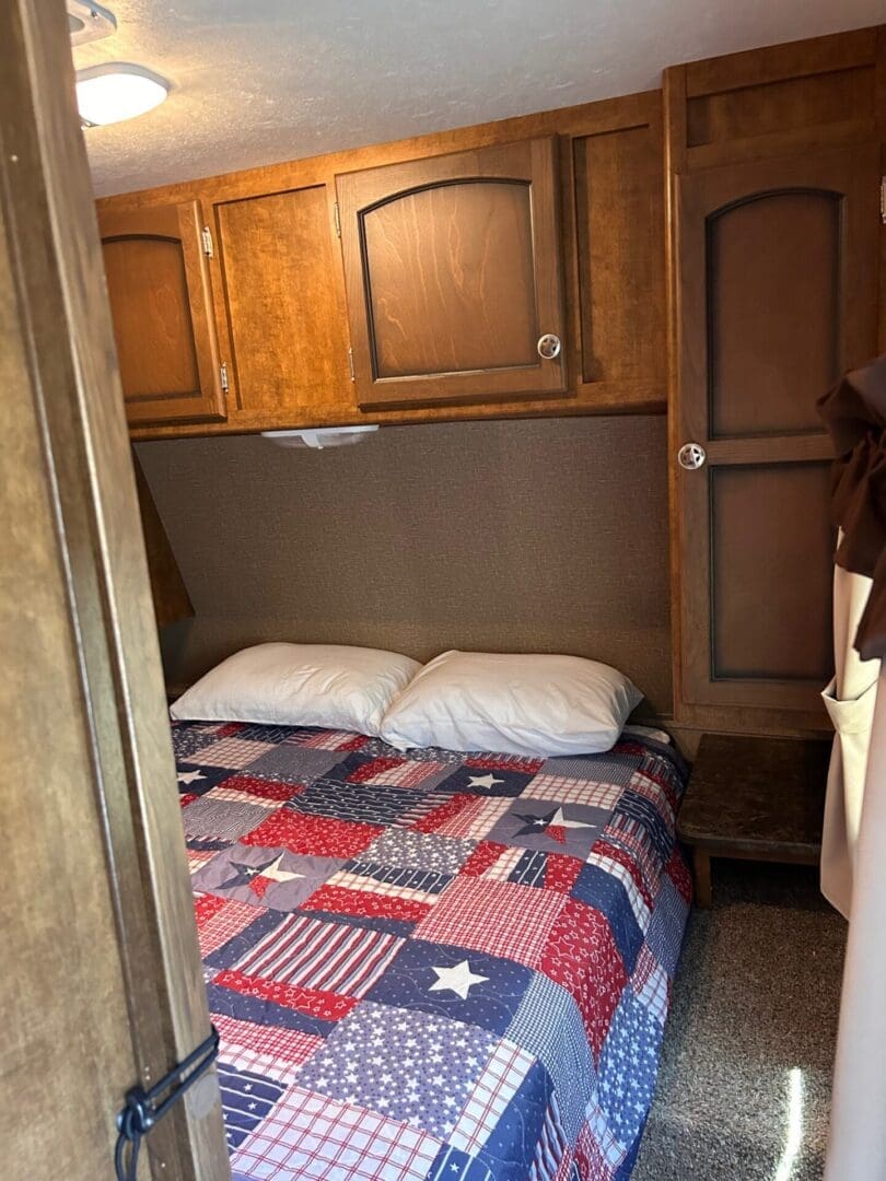 Interior of a camper with a bed covered in a patriotic quilt and overhead wooden cabinets, lit by natural light.