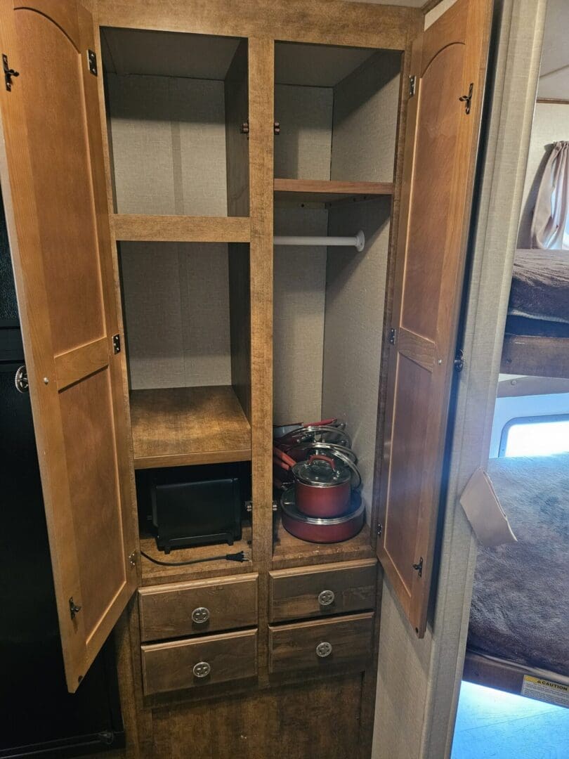 Open wooden wardrobe in an rv with empty shelves, two drawers, one containing a red kettle and black pot, and doors partially open.