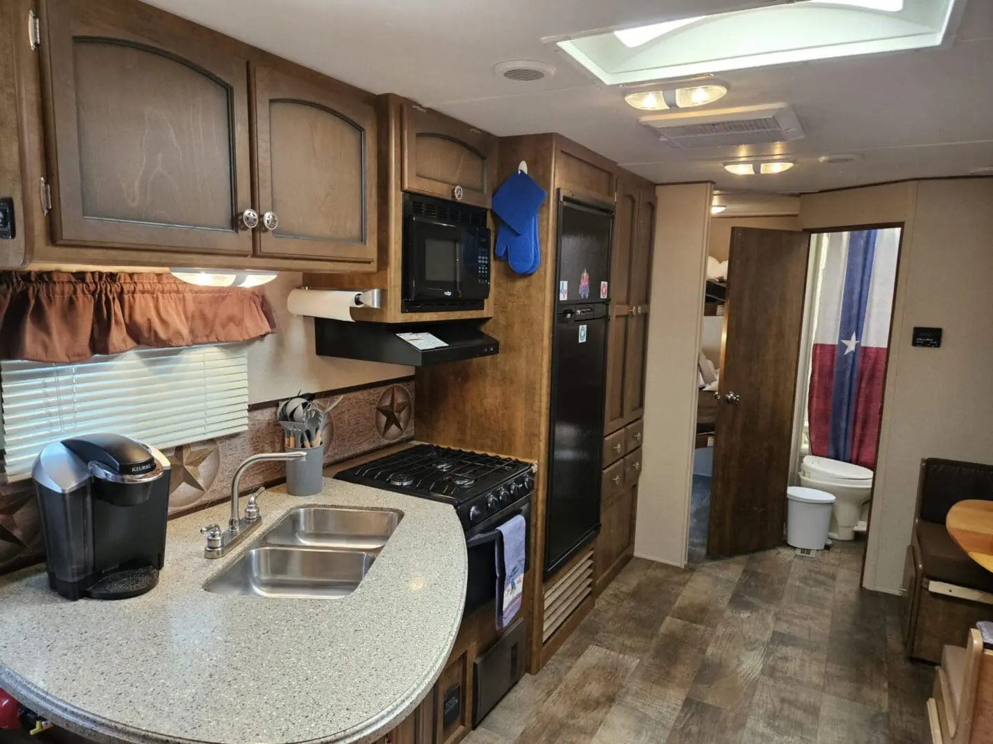 Interior of a motorhome showing a kitchen with a sink, stove, oven, microwave, refrigerator, and an open bathroom door at the end.