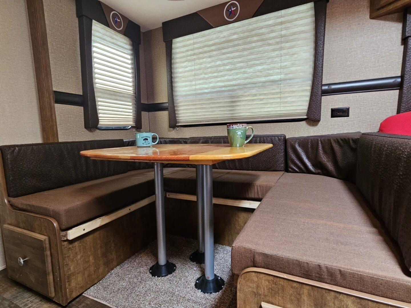 Interior of a camper with a u-shaped dinette, wooden table, two mugs, and surrounding cushioned seats.