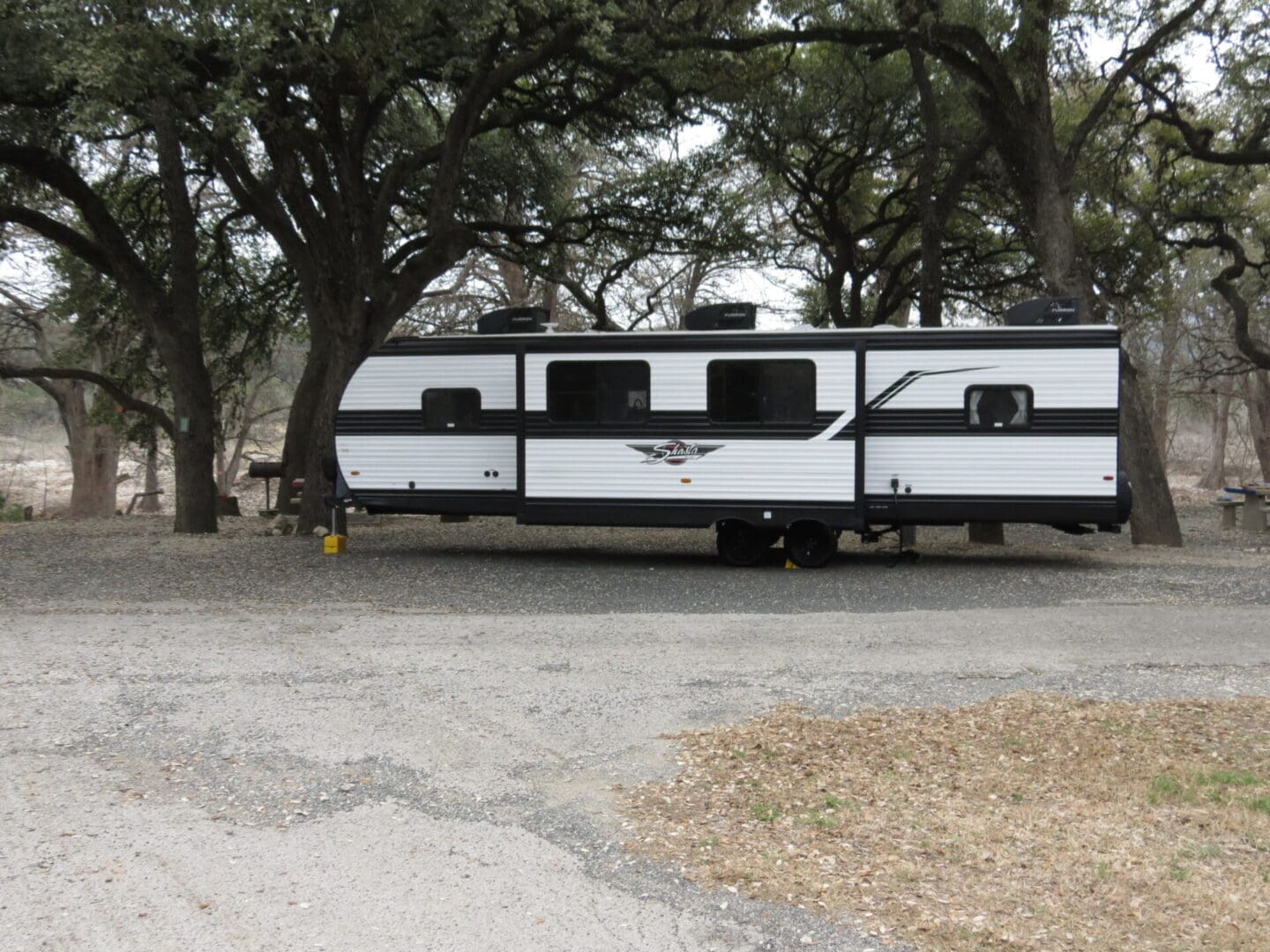 Large travel trailer parked under oak trees on a gravel lot.