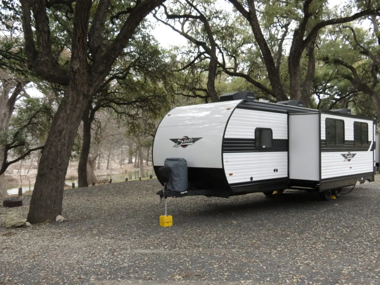 A white travel trailer parked under oak trees on a gravel lot.