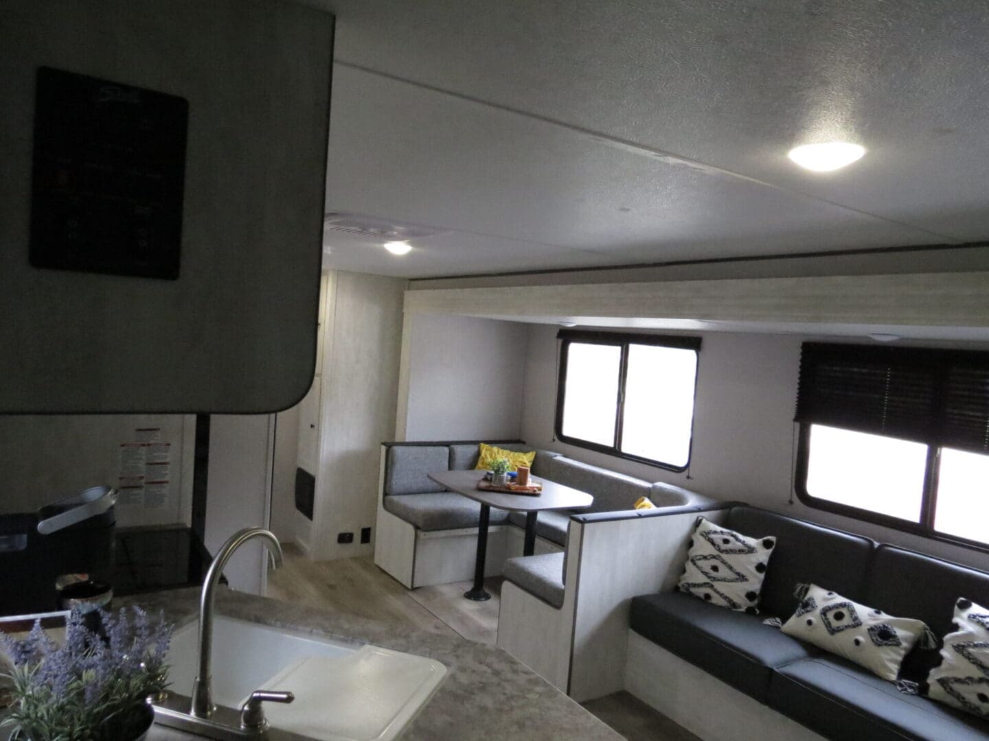 Interior of a modern rv featuring a kitchen area with a sink in the foreground and a dining area with benches and a table in the background, all under bright ceiling lights.