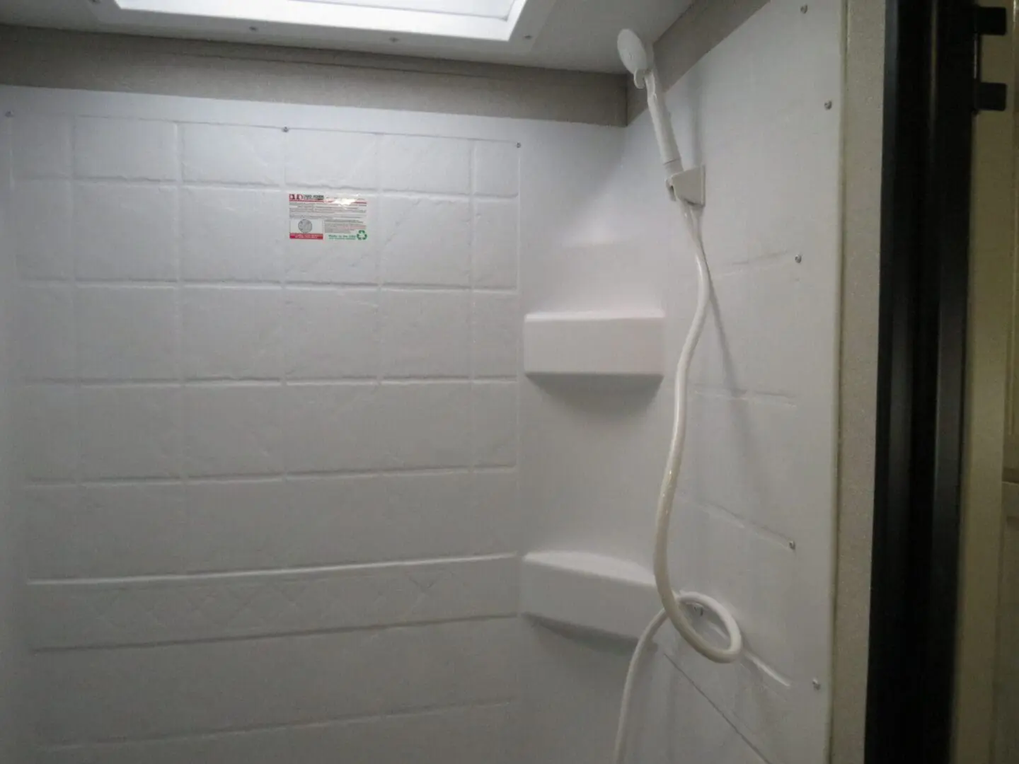 Interior of a white shower cabin with a handheld showerhead hanging on the wall, and a small shelf mounted below a cautionary sign.