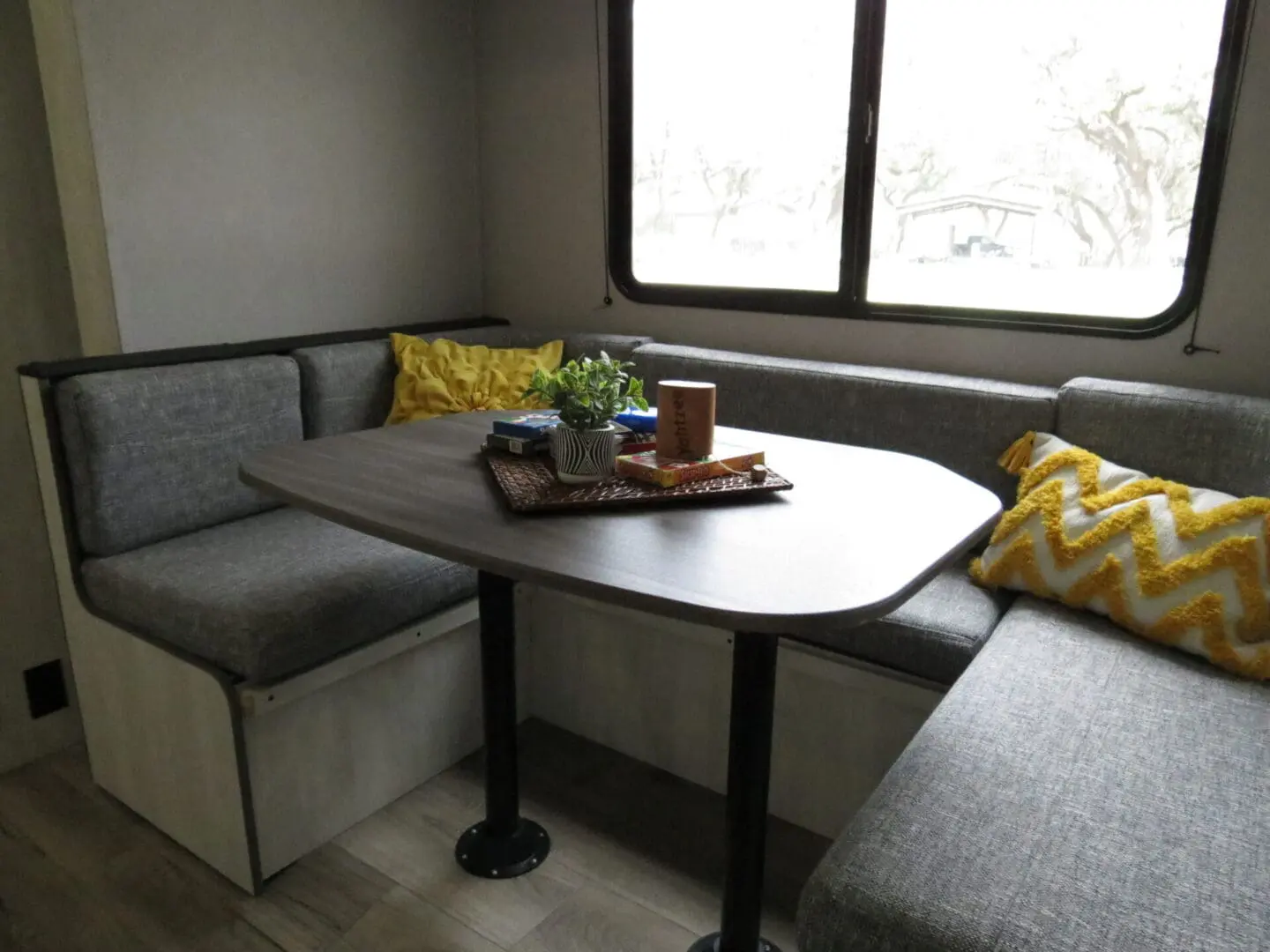 Cozy rv interior with a u-shaped dinette set, featuring grey cushions, a dark table, and decorative items like a plant and books.