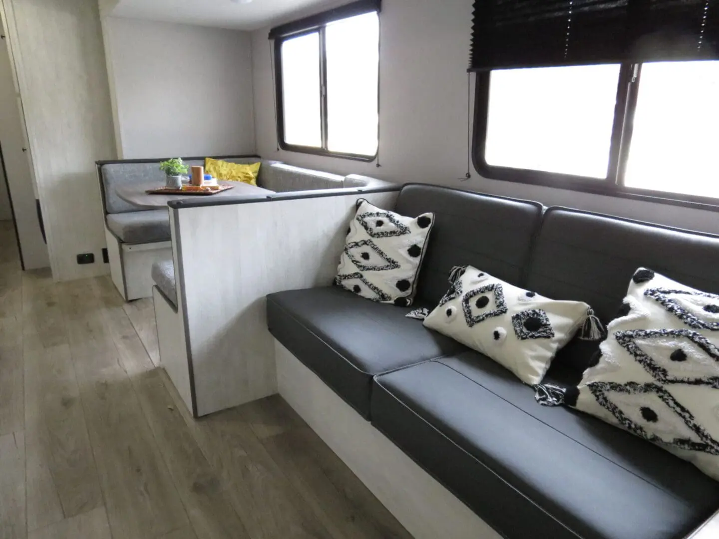 Interior of a modern rv featuring a dining area with a booth-style table and a living area with a dark couch and decorative pillows.