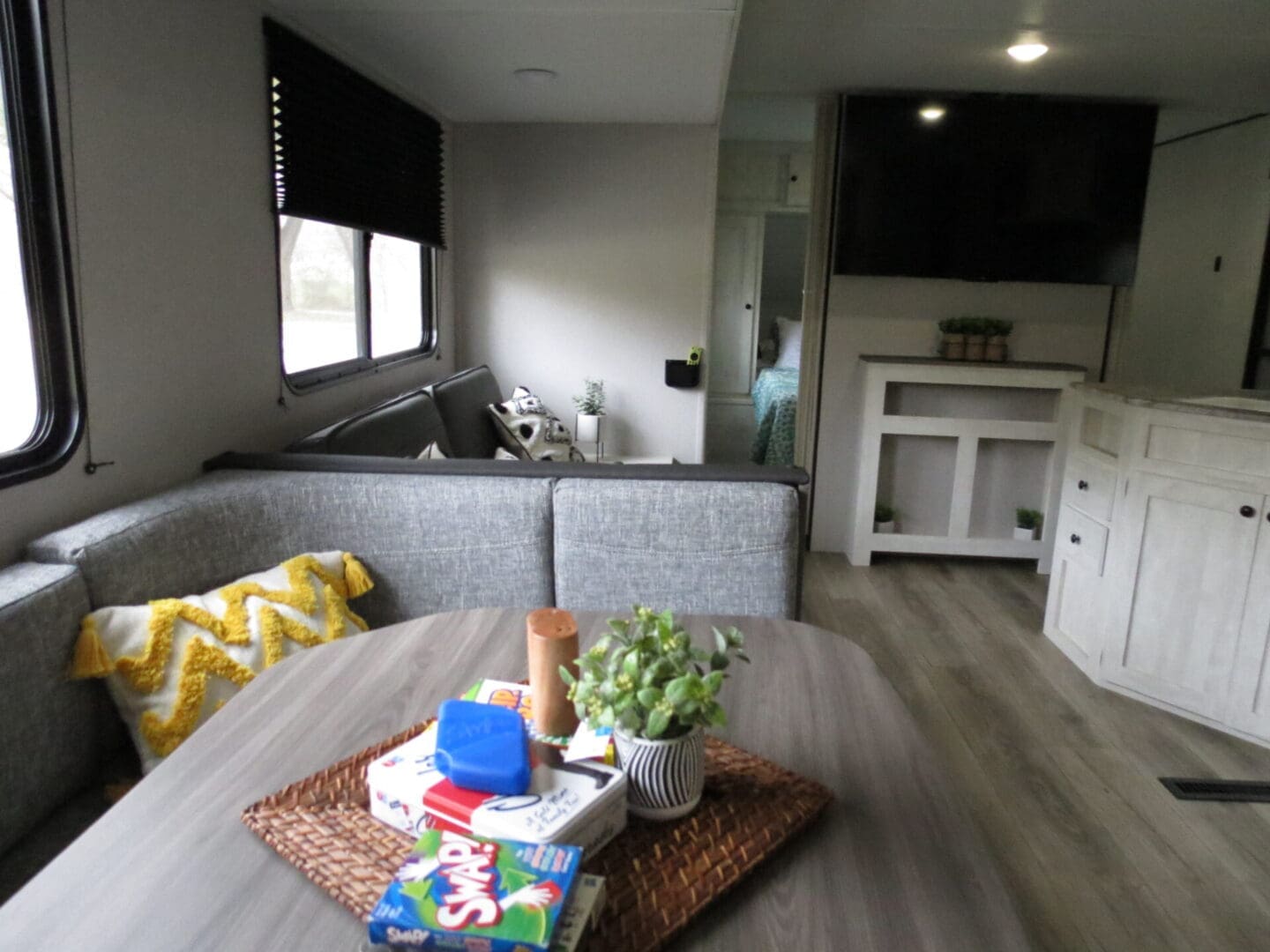 Interior of a modern rv featuring a cozy sitting area with a gray sofa, patterned pillows, a small dining table, and a flat-screen tv mounted on a white cabinet.