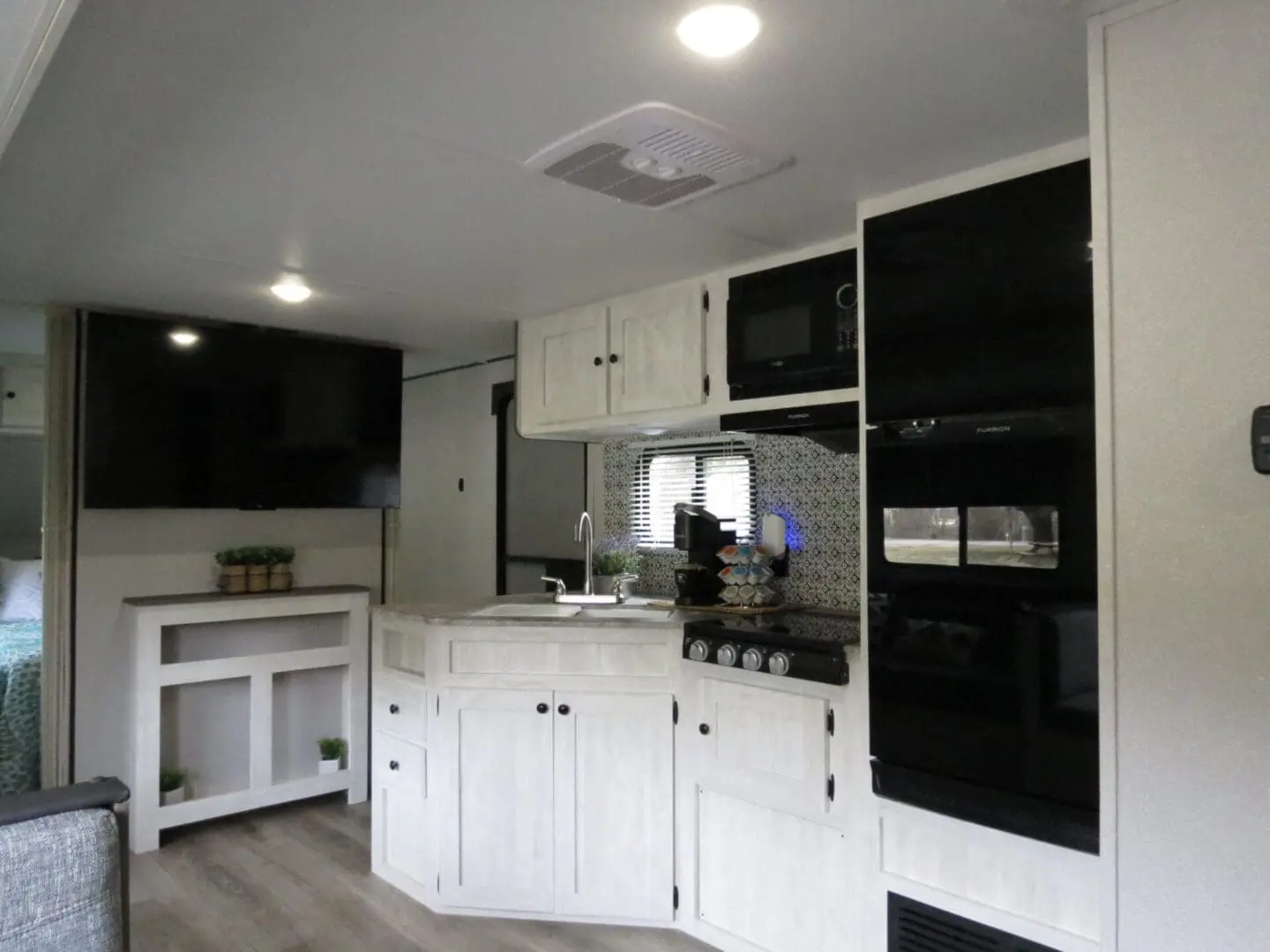 Interior of a modern rv kitchen featuring white cabinets, a microwave, stove, and a small dining area, with a television mounted on the wall.