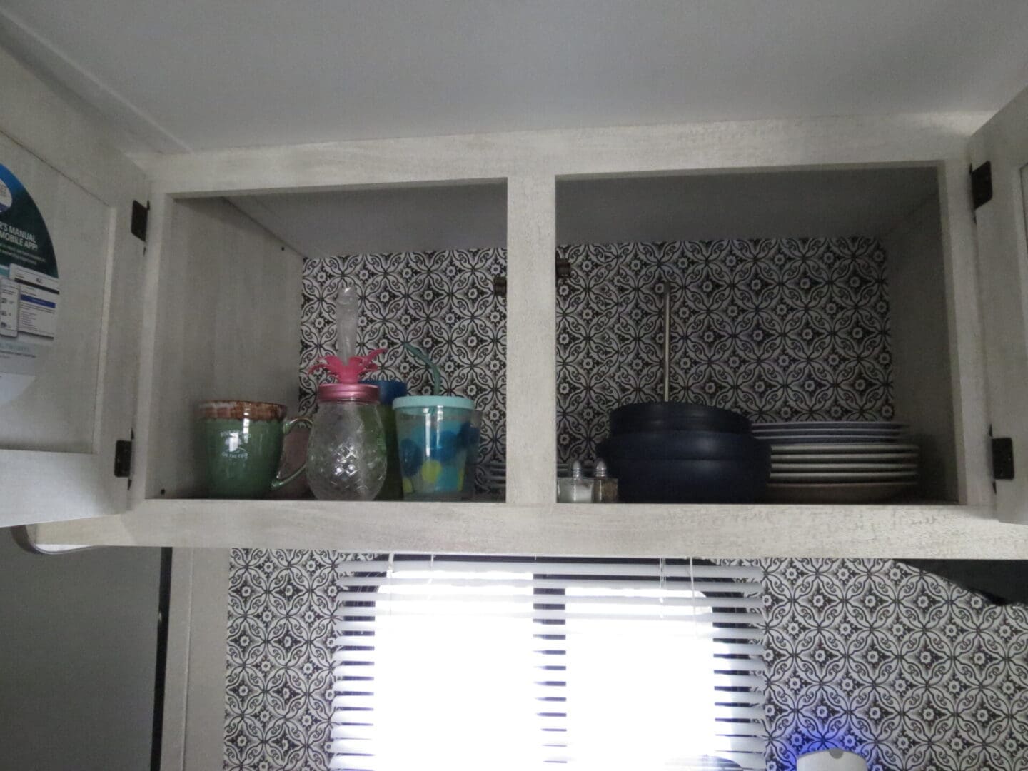 Open kitchen cabinet with patterned background containing assorted jars, a pink water bottle, and piles of plates and cooking pots.