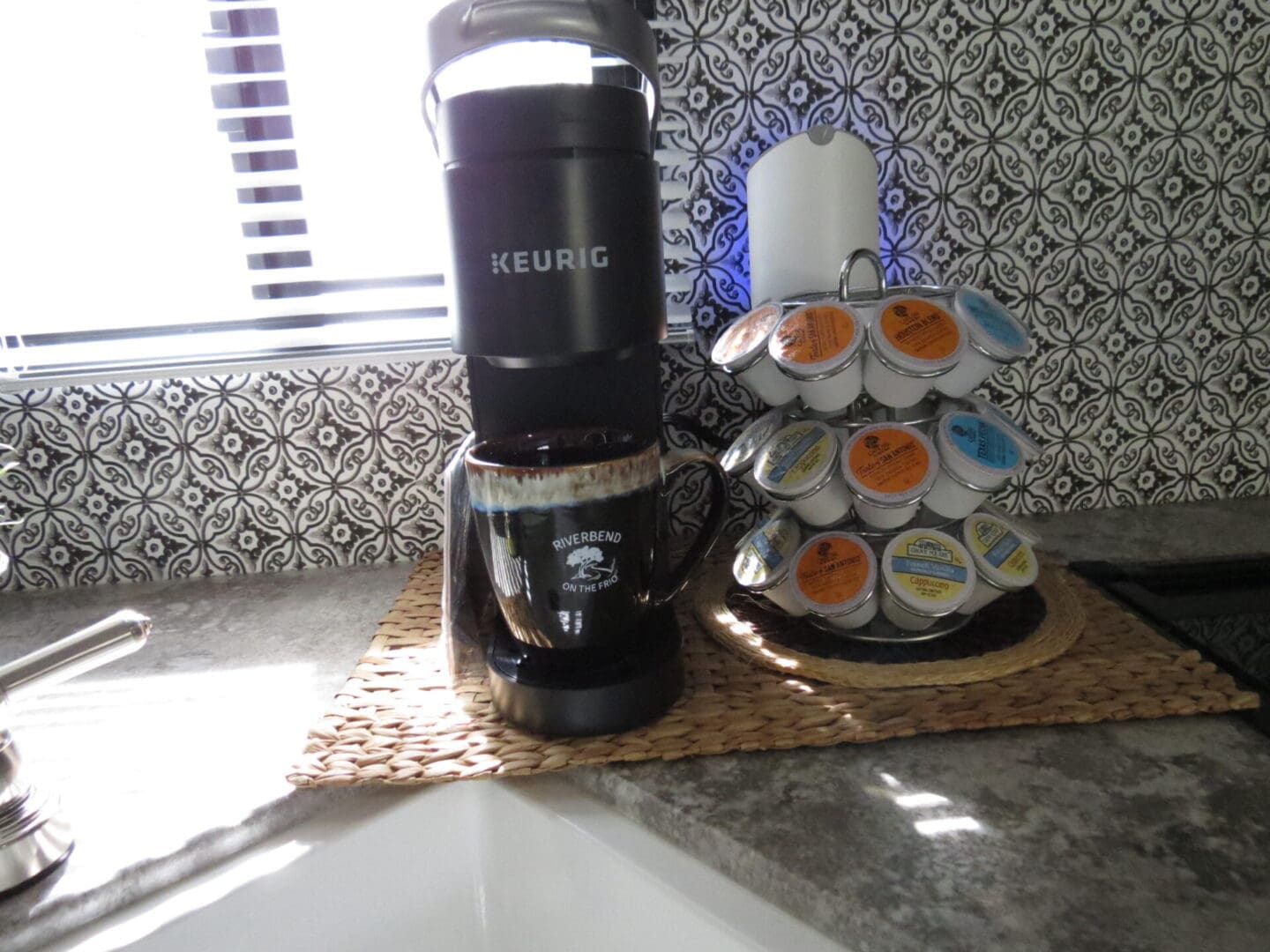 A keurig coffee maker dispensing coffee into a mug beside a carousel of coffee pods on a kitchen counter with a patterned backsplash.