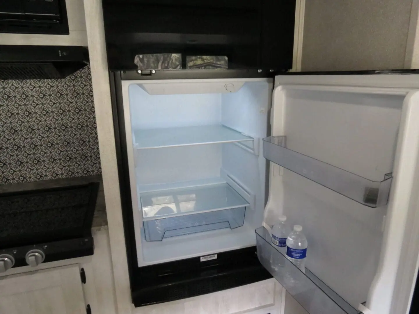 An open, nearly empty refrigerator inside an rv kitchen with two bottles of water on the bottom shelf and a tiled backsplash.