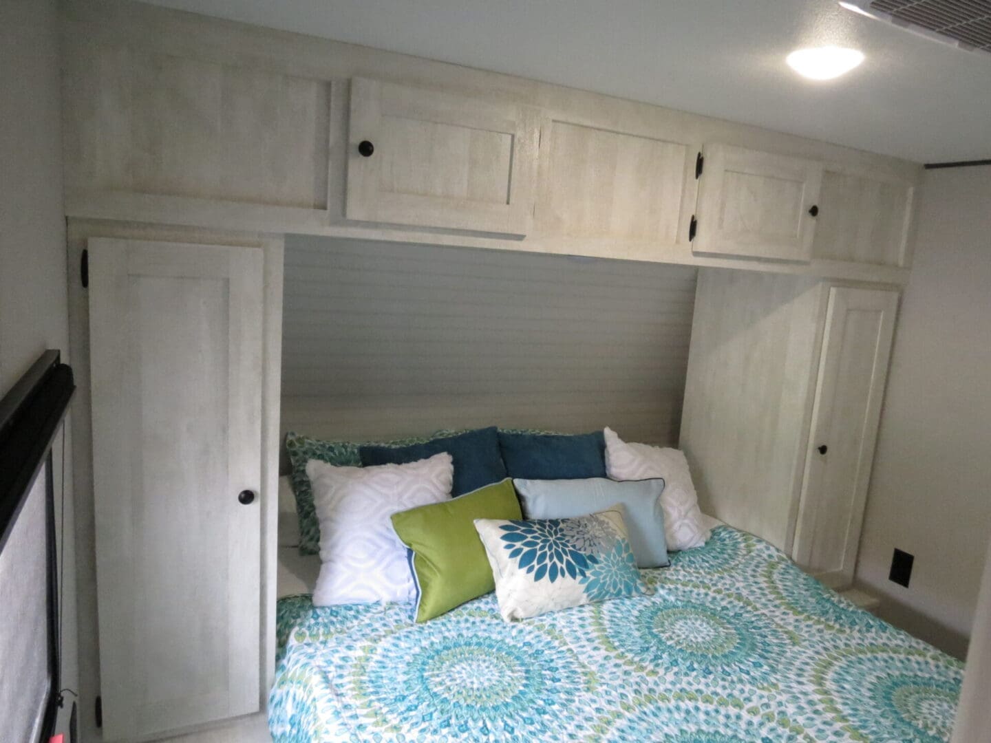 A small bedroom featuring a bed with a blue and green quilt, flanked by closed white wooden cabinets and overhead storage.