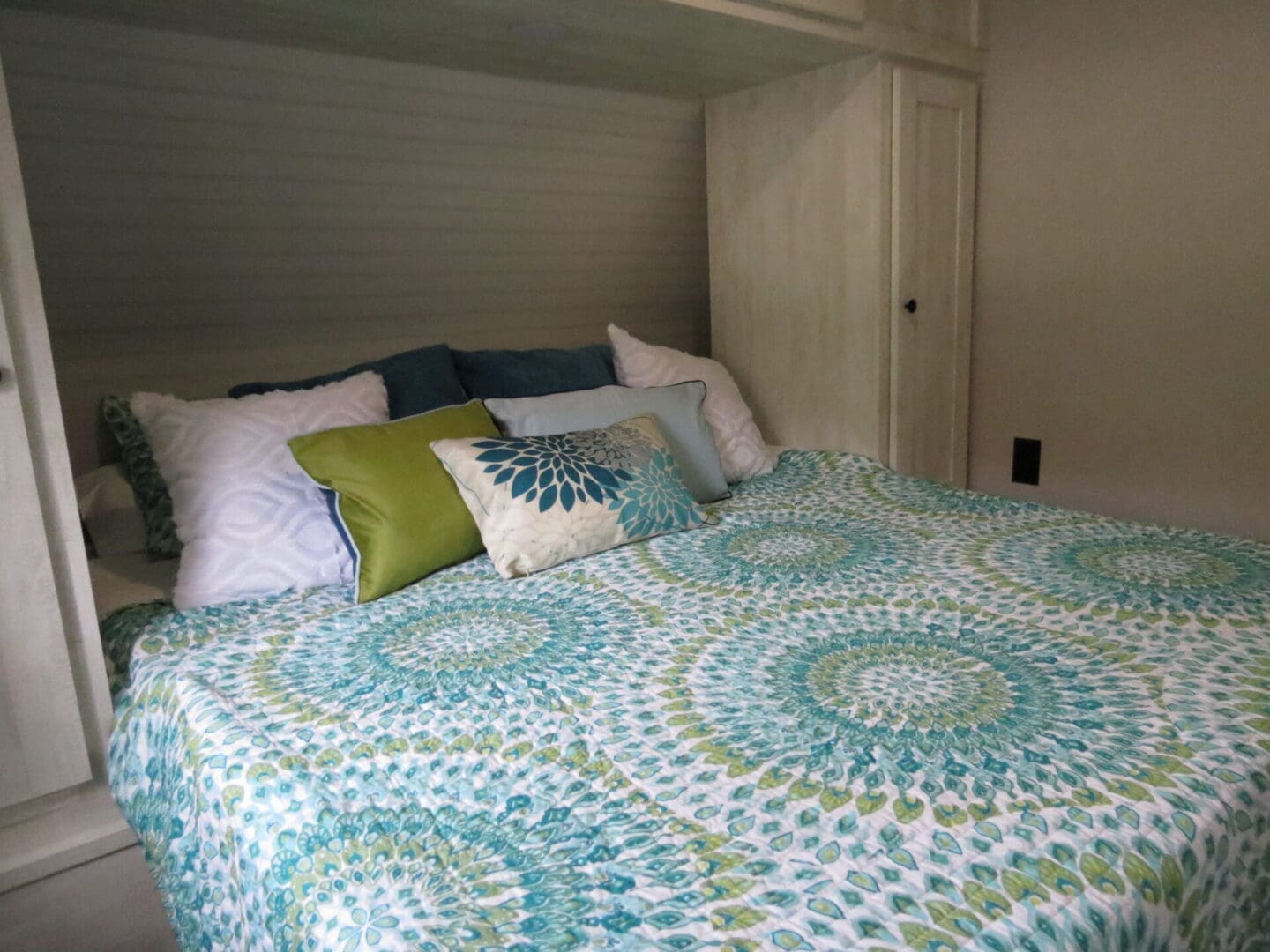 A small bedroom with a neatly made bed covered in a blue and green floral quilt, surrounded by white wooden walls and a cabinet.