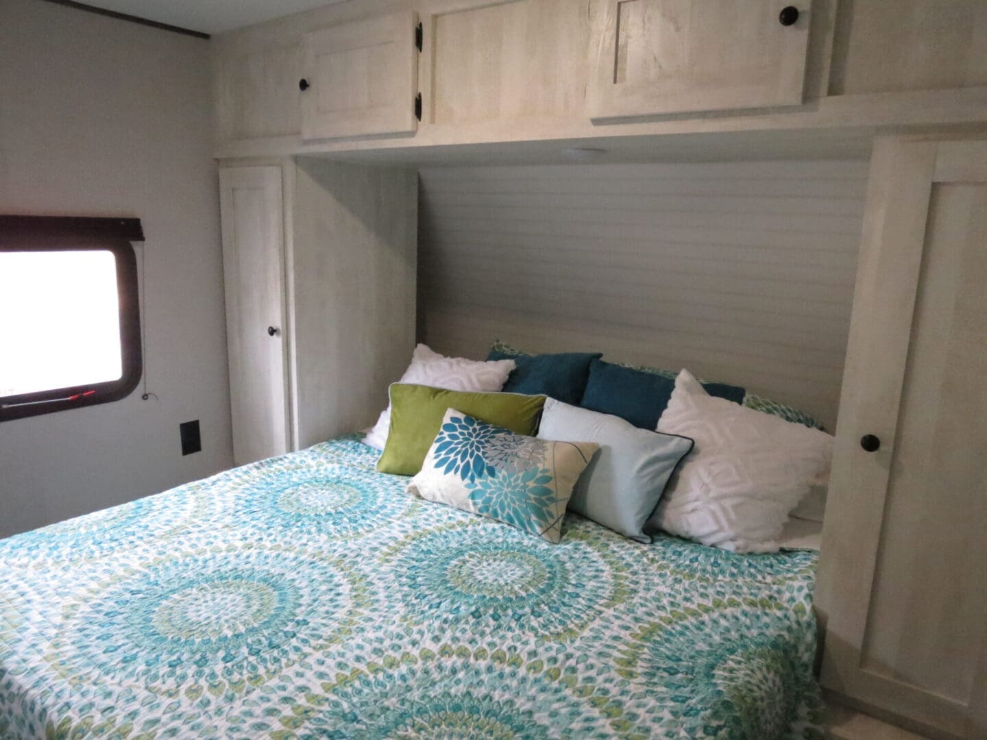 A cozy bedroom in an rv featuring a bed with a patterned teal and white comforter, multiple colorful pillows, and wooden overhead cabinets.