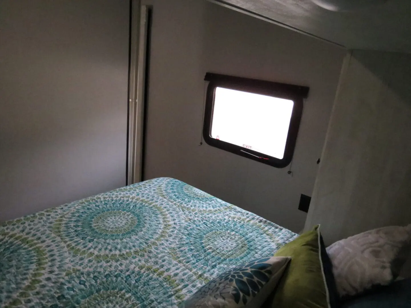 Interior of a small bedroom in an rv with a bed covered in a blue and green quilt near a single window with the blinds up.