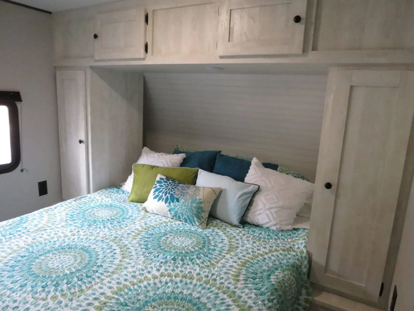 A cozy rv bedroom featuring a bed with a decorative teal and green quilt, multiple pillows, and overhead wooden cabinets.