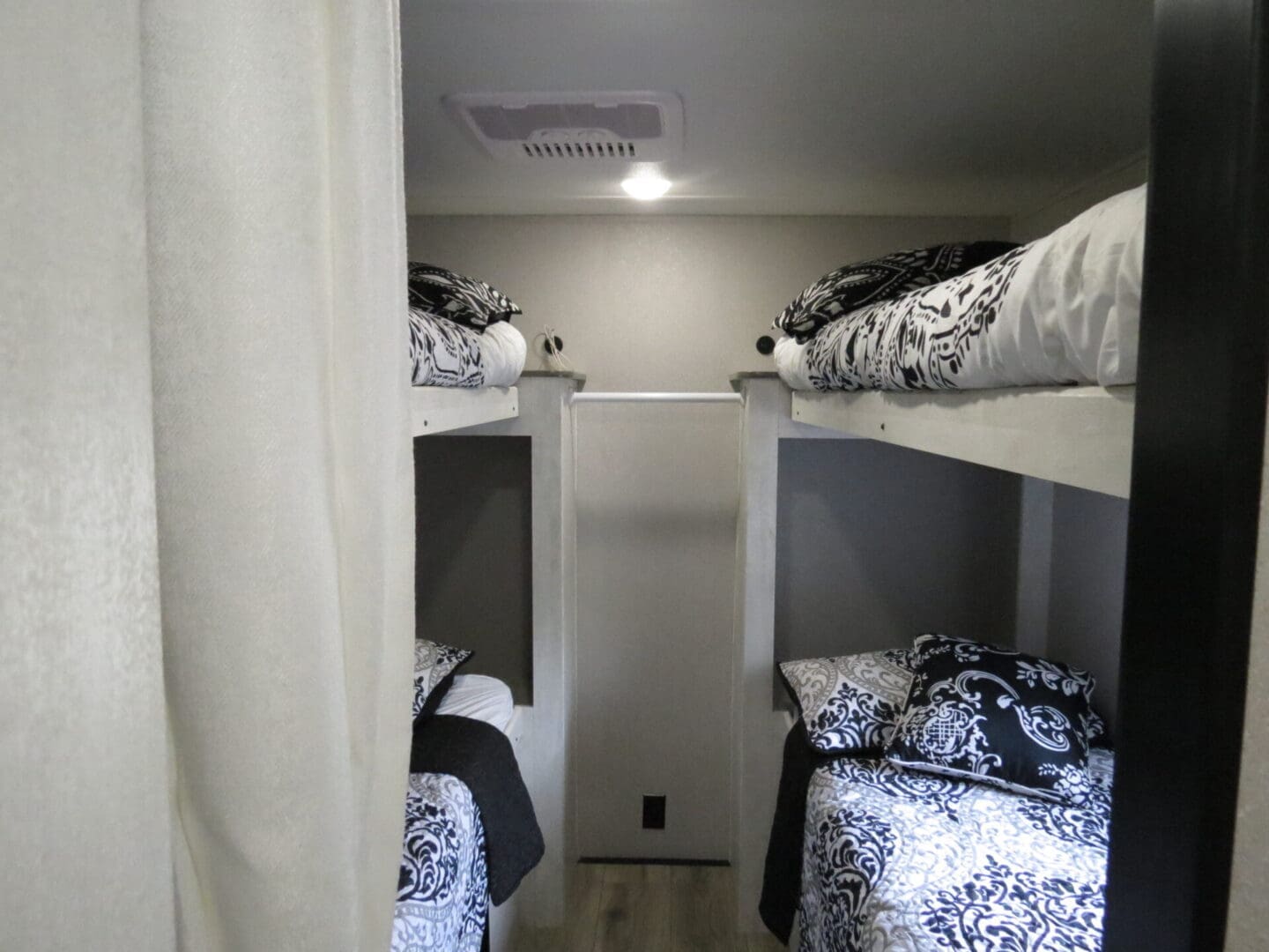 Interior of a caravan showing two sets of bunk beds with black and white bedding, curtains partially drawn, providing privacy.