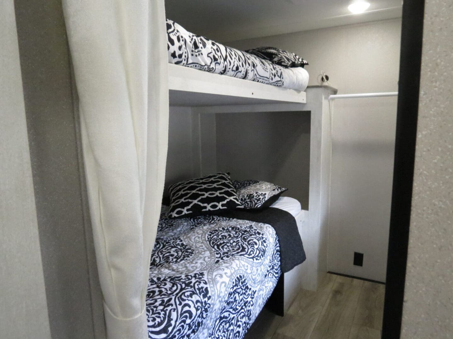 A tidy bunk bed setup in a room, with both beds covered in black and white patterned bedding and curtains partially drawn around the lower bed.