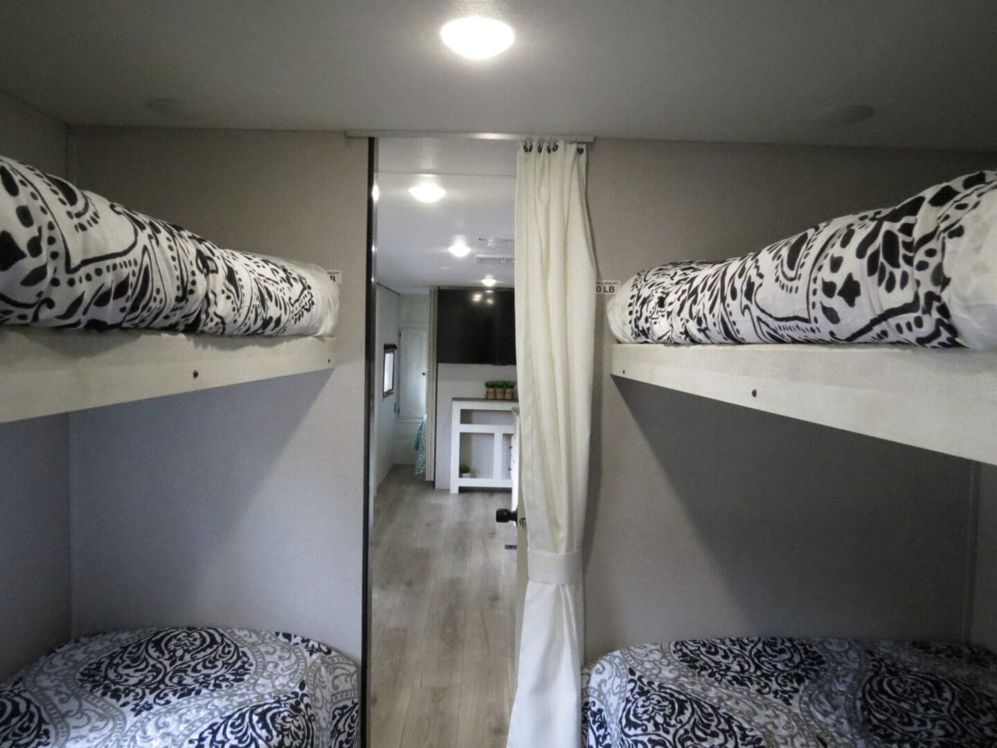 Interior of a recreational vehicle showing two bunk beds with patterned bedding, and a view into a kitchen area beyond a curtain.