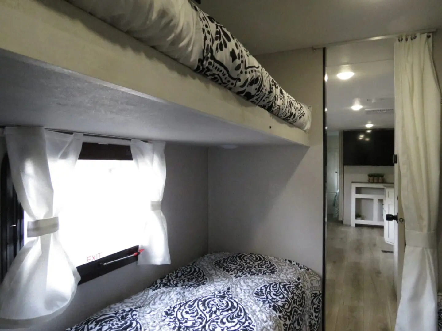 Interior of a modern camper with a bunk bed setup; the lower bed is dressed with a black and white patterned cover, and there's a window with curtains beside it.