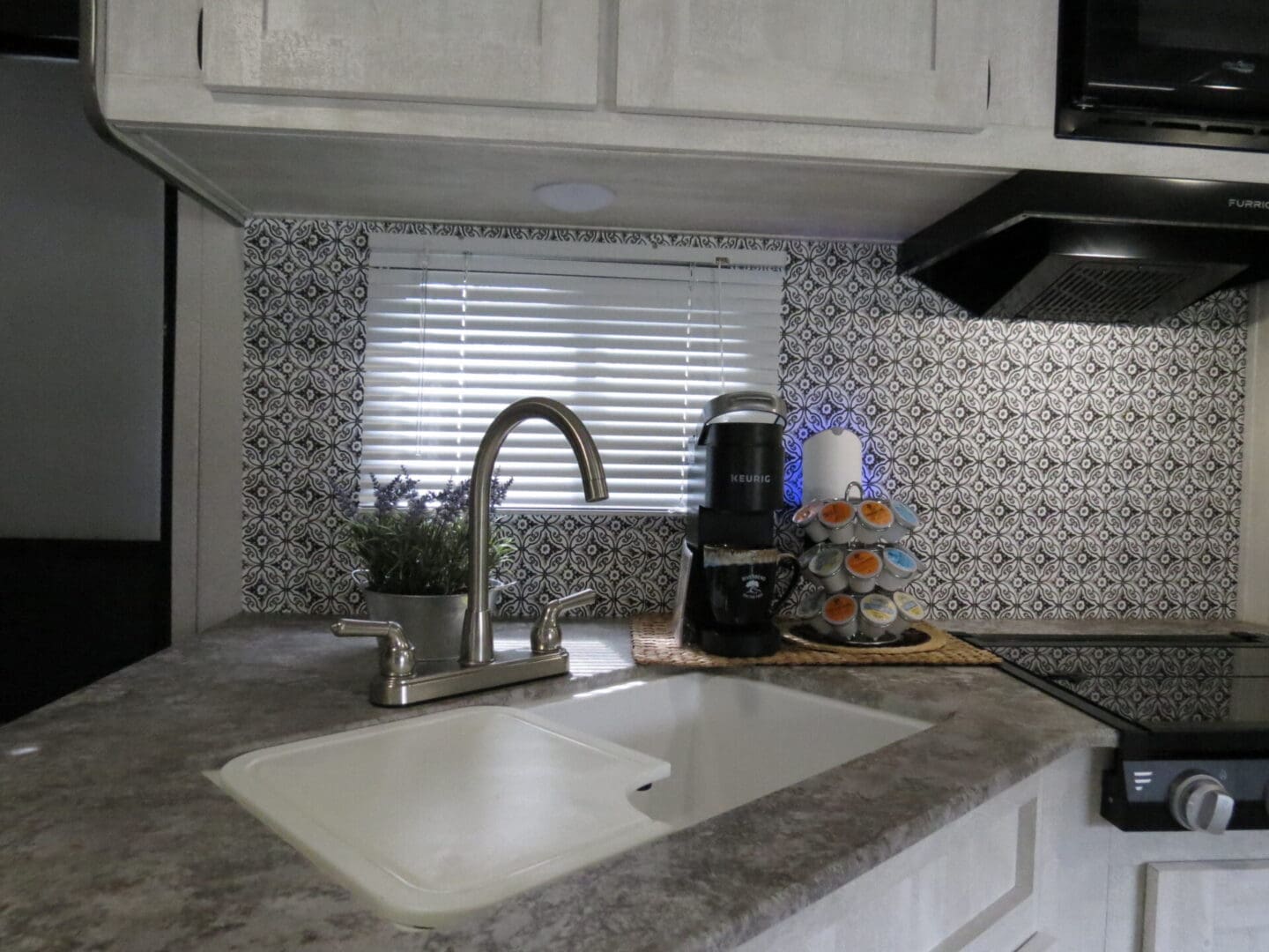 Modern kitchen corner featuring a sink, faucet, and a coffee maker set against a decorative backsplash, with a venetian blind-covered window above the sink.