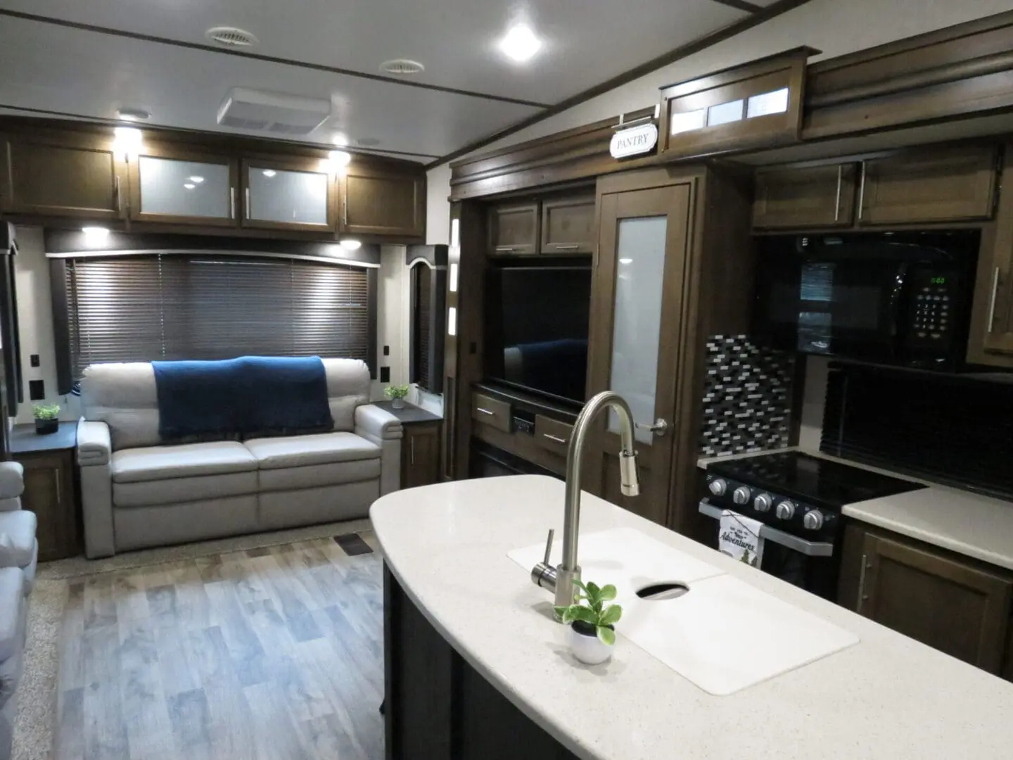 Interior of a modern rv featuring a kitchen with sink on a countertop, a living area with a blue sofa, a wooden cabinet with a tv, and muted lighting.