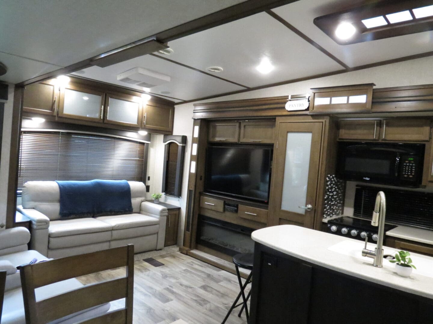 Interior of a modern rv featuring a kitchen area with a microwave and sink, a living area with a sofa and tv, and wooden cabinets throughout.