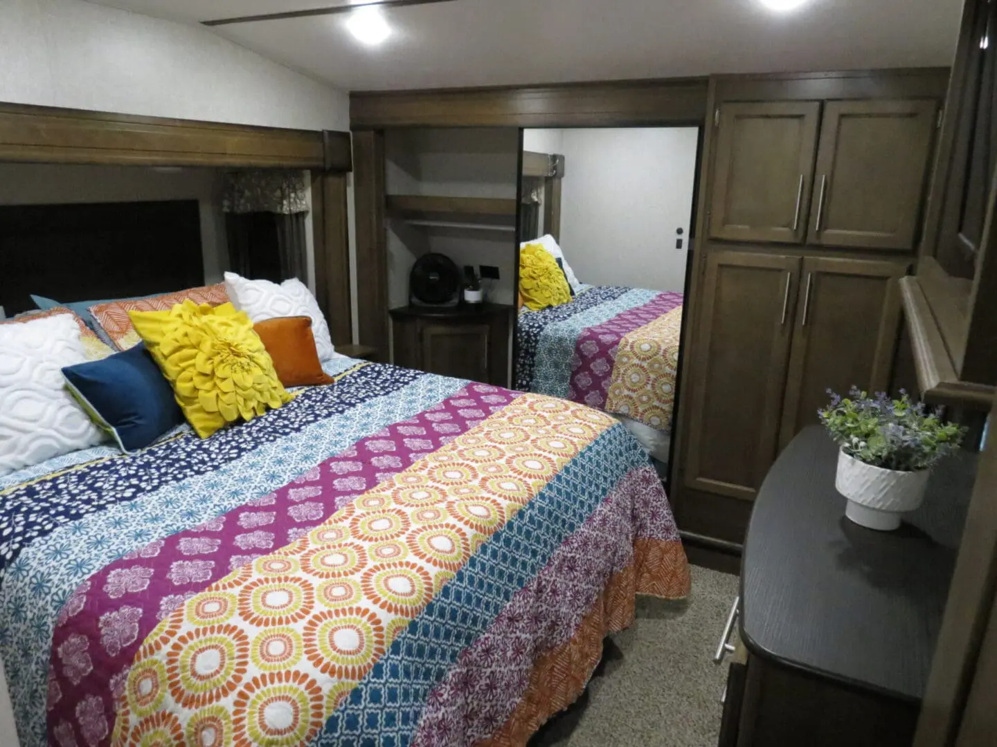 Interior of a cozy rv bedroom featuring two single beds with colorful quilts and decorative pillows, flanked by wooden cabinets and a small bedside table with a plant.