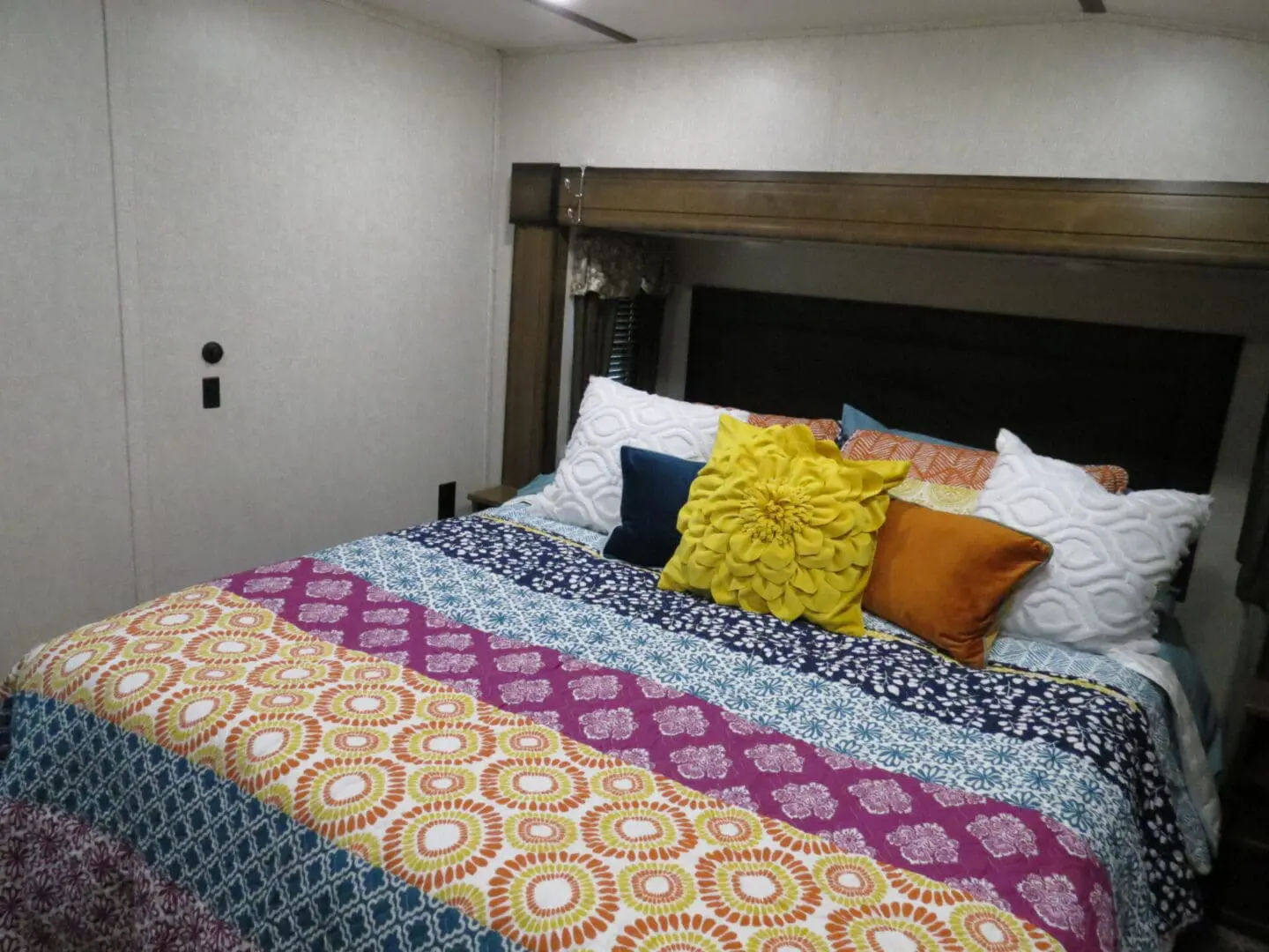 Interior of an rv bedroom featuring a quilted bed with multiple colorful pillows and wooden headboard.