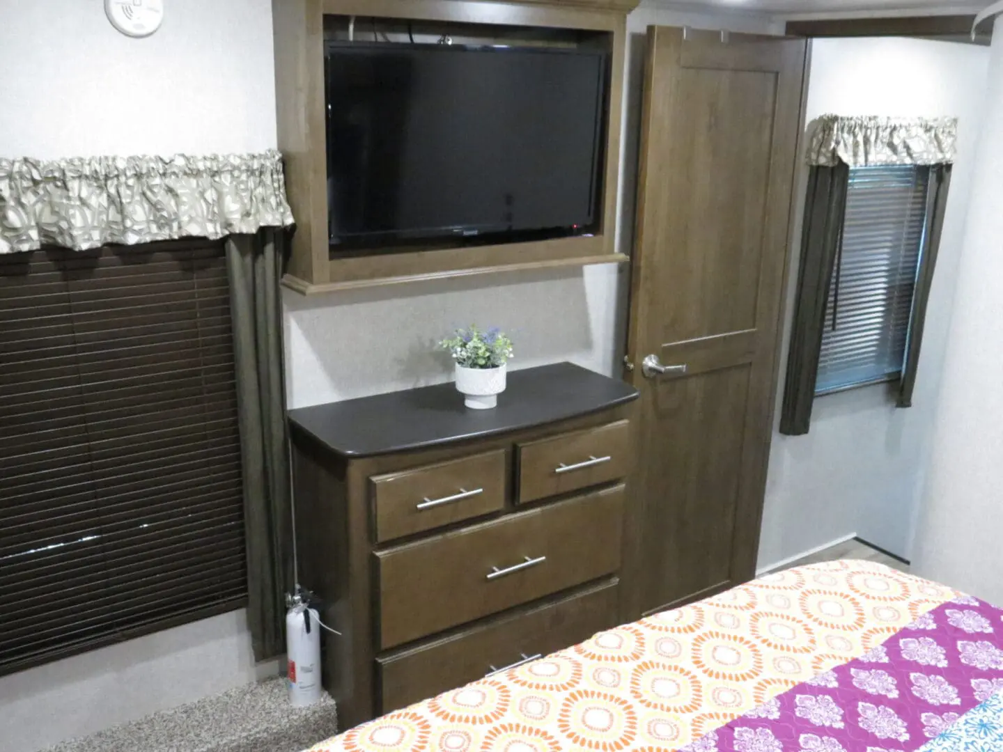 Interior of a recreational vehicle with a floral bedspread, wood cabinets, and a mounted tv above a dresser with a potted plant.