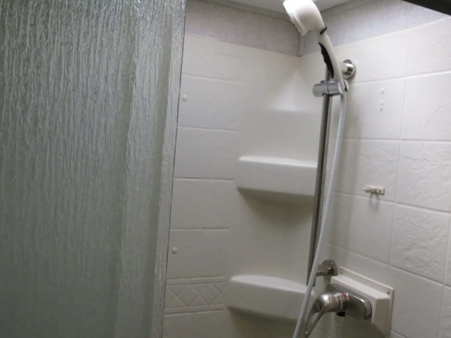 A small bathroom corner featuring a shower with white tiled walls, built-in shelves, and a flexible hose showerhead.