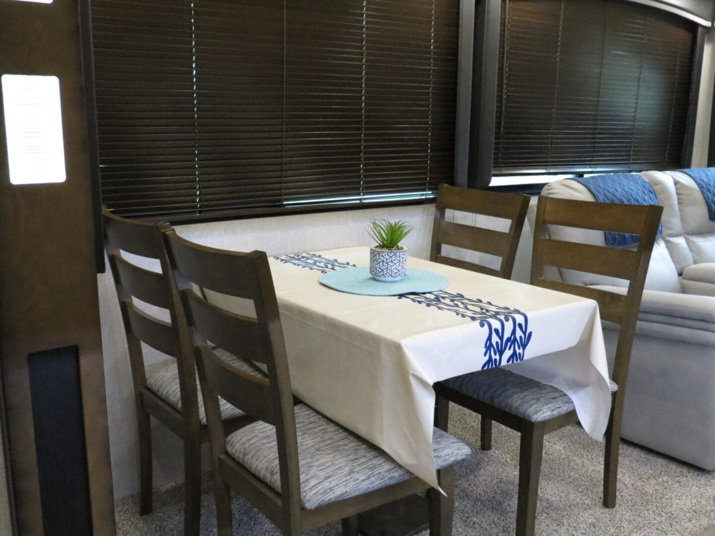 A neatly arranged dining area inside an rv, featuring a table with four chairs, set with a blue centerpiece and a small potted plant, flanked by large windows with brown blinds.