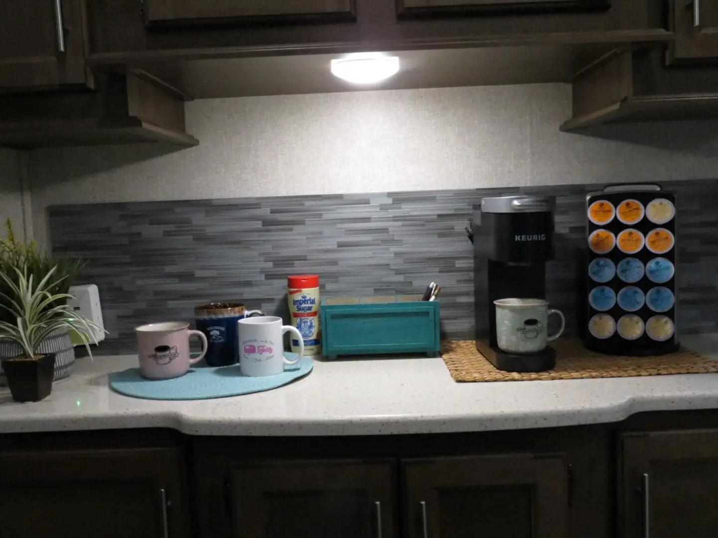 Kitchen counter featuring a coffee maker, cups, tea and coffee pods, and a small potted plant under cabinet lighting.