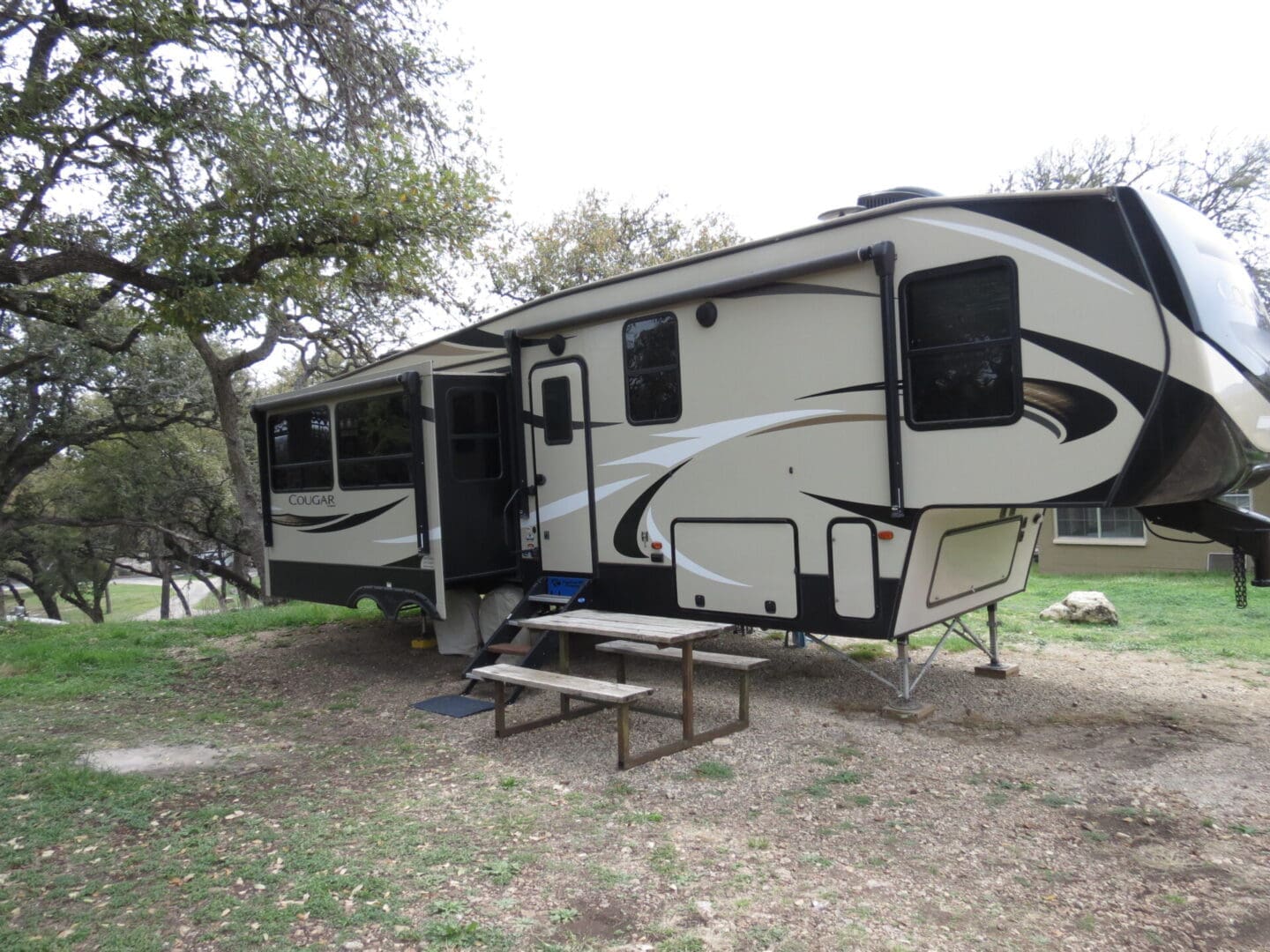 A modern camper trailer parked in a grassy area surrounded by trees, featuring multiple slide-outs and a picnic table nearby.