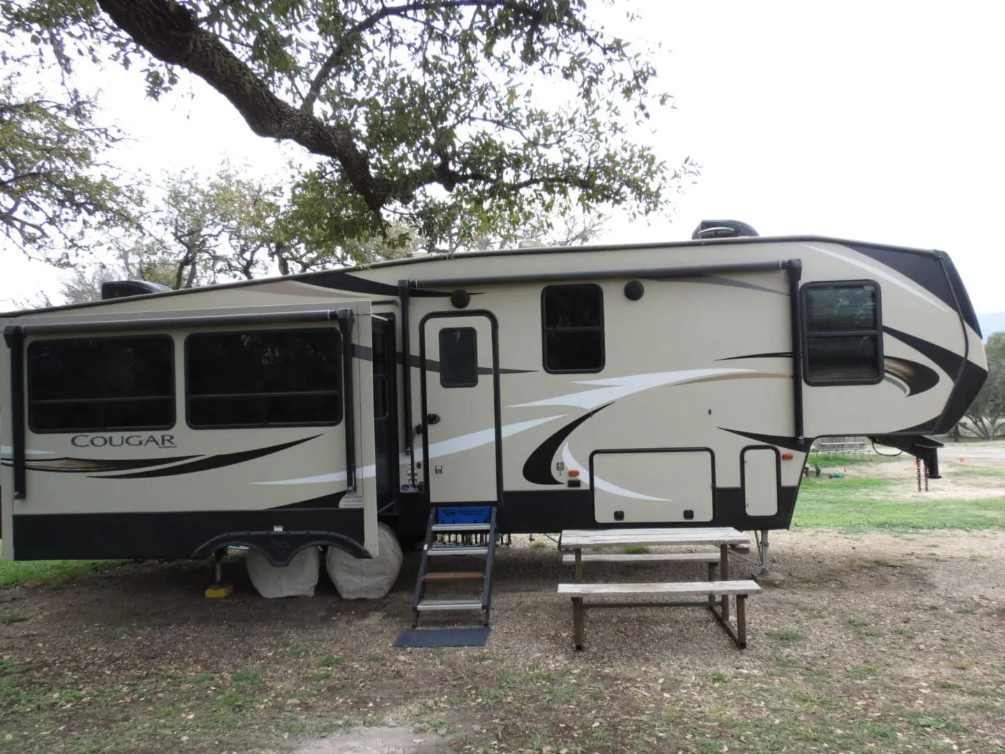 A large cougar fifth-wheel rv parked outdoors with extended slide-outs and a picnic table nearby under leafy trees.